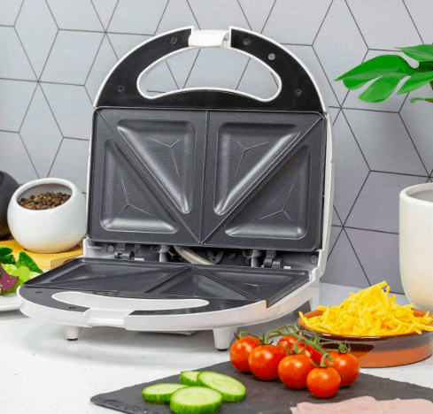 toastie maker opened on a kitchen counter with sliced vegetables and a plate of cheese on the side.