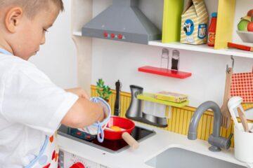 little boy putting his hand in a white and blue oven mitt, with a toy kitchen in front of him, including a stove, sink and shelves with toy bottle of milk and ketchup