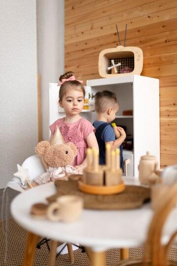 little girl and a toddler playing pretend tea party with wooden cake toy and wooden teacups and their teddy bear, the girl is looking at the table where the wooden cake is and the toddler behind her looking at the toy fridge