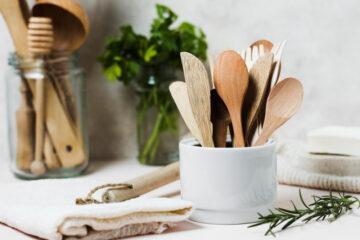 Wooden cutlery is displayed inside a porcelain bowl, you can see blurry wooden kitchen accessories in the background as well as celery in a glass jar