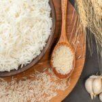 Superior view of a bowl full of cooked rice, there is a spoon with uncooked rice grains beside the bowl and also grains around the plate.