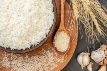 Superior view of a bowl full of cooked rice, there is a spoon with uncooked rice grains beside the bowl and also grains around the plate.