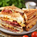 The image displays a side view of three tall sandwiches made with a sandwich maker cut in half resting on a plate; the sandwiches are skewered with a toothpick