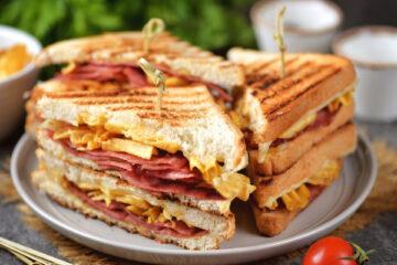 The image displays a side view of three tall sandwiches made with a sandwich maker cut in half resting on a plate; the sandwiches are skewered with a toothpick