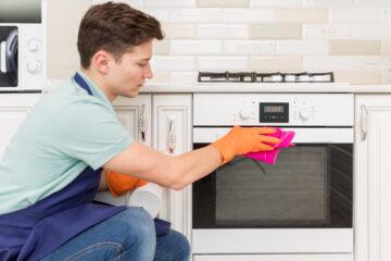 Young man using gloves and oven cleaning products to clean the oven in a white kitchen