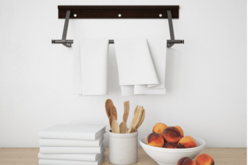 Below the tea towel holder fixed on the wall, there is a table with kitchen utensils, a bowl of fruit, and towels.