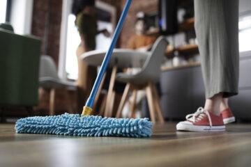 a display of a person cleaning the floor tiles with a blue mop, a blurred background of another person in the room standing in a kitchen