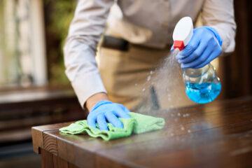 A lady wearing gloves and cleaning the table with a cleaner spray and a towel