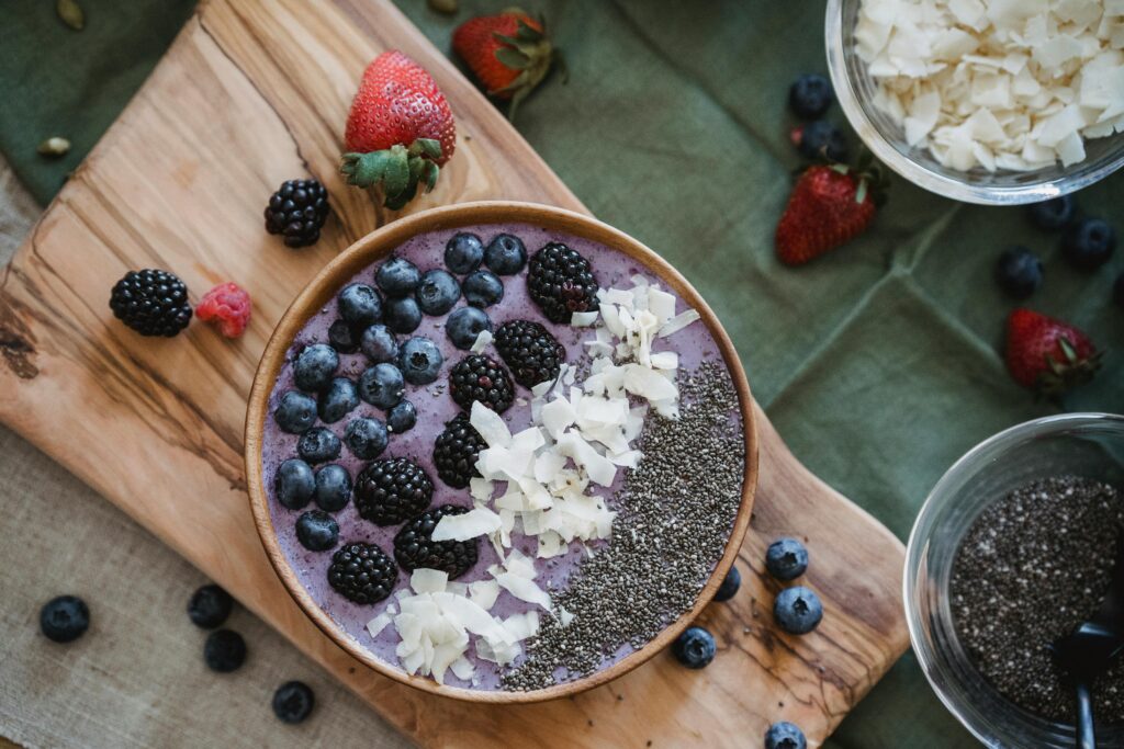 A healthy berry smoothie bowl topped with fresh fruits, seeds, and coconut shavings on a wooden board.